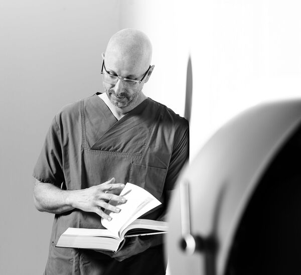 Schwarz-weiß-Foto von Axel Hillebrandt in medizinischer Kleidung, der an einer Wand lehnt und in einem aufgeschlagenen Buch liest.
