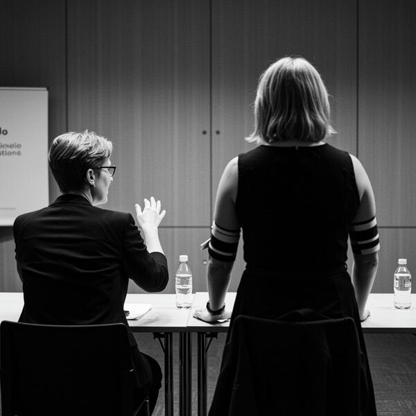 Schwarzweiß-KI-Fotografie von zwei Personen an einem Tisch in einem Konferenzraum, eine Person sitzend gestikulierend, die andere stehend, mit Wasserflaschen auf dem Tisch.